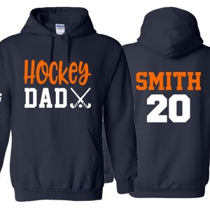 May include: Two navy blue hooded sweatshirts. The left sweatshirt has the text "Hockey Dad" with crossed hockey sticks in orange and white. The right sweatshirt has the text "SMITH 20" in white and orange.