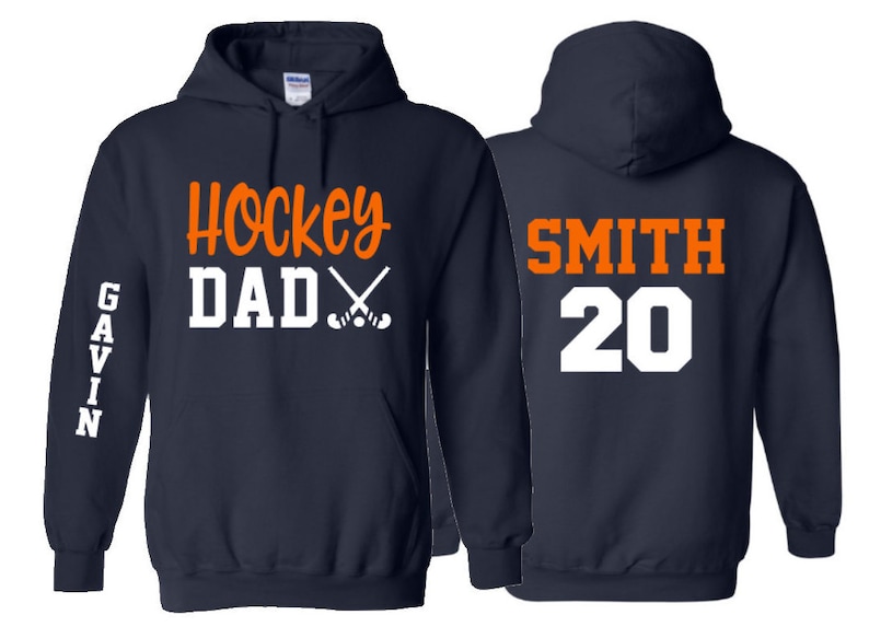 May include: Two navy blue hooded sweatshirts. The left sweatshirt has the text "Hockey Dad" with crossed hockey sticks in orange and white. The right sweatshirt has the text "SMITH 20" in white and orange.