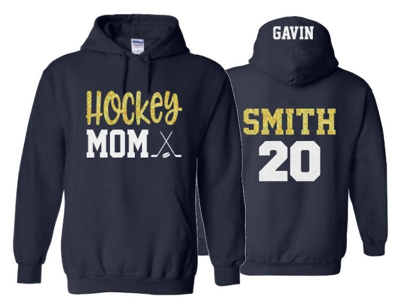 May include: Navy blue hoodie with "Hockey Mom" in gold glitter and white text, plus hockey sticks. The back features "GAVIN" above "SMITH 20" in gold glitter and white. A comfortable, sporty apparel item.