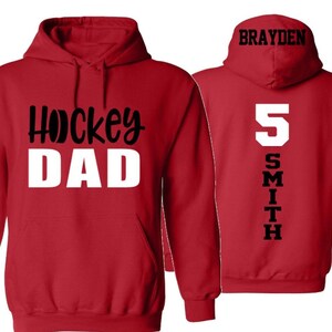 Op de afbeelding: Rood hoodie met de tekst "Hockey Dad" in zwart en wit op de voorkant en de tekst "Brayden Smith 5" in zwart op de achterkant.