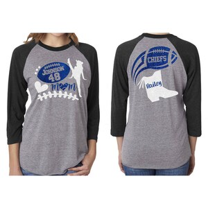 May include: A gray and black baseball tee shirt with a white silhouette of a cheerleader kicking a football. The front of the shirt has a football with the text "Johnson 48" and a heart with the word "Mom". The back of the shirt has a football with the text "Chiefs" and a white silhouette of a cheerleader's boot with the name "Hailey".