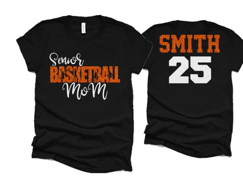 K&ouml;nnte beinhalten: Zwei schwarze T-Shirts. Ein Shirt sagt "Senior Basketball Mom" in orangefarbenem Glitzer mit schwarzen Basketballspielern. Das andere Shirt sagt "SMITH 25" in wei&szlig;em Glitzer.