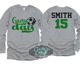 May include: Gray long-sleeve shirt with "Game Day" in green script and a soccer ball graphic. The back displays "SMITH" in black and the number "15" in green. A logo is visible at the bottom.