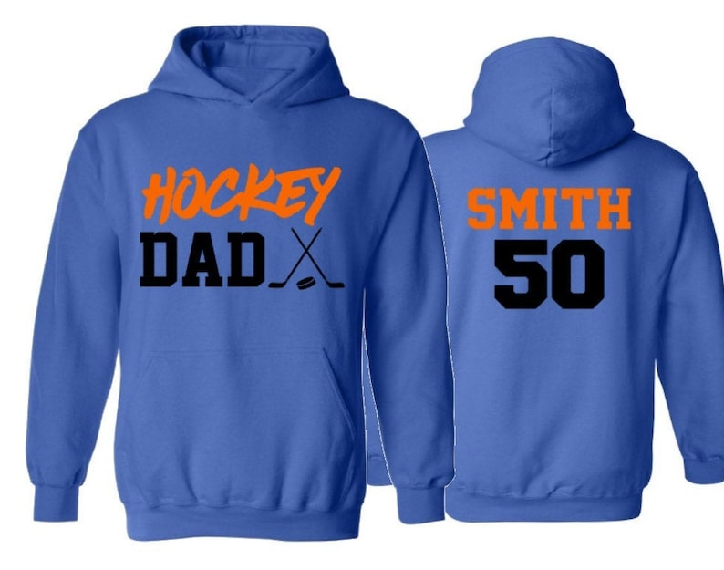 Pode incluir: Dois casacos de moletom com capuz azuis. A frente de um casaco tem o texto "HOCKEY DAD" em laranja com um taco de h&oacute;quei e um disco em preto. As costas do outro casaco tem o texto "SMITH 50" em laranja e preto.