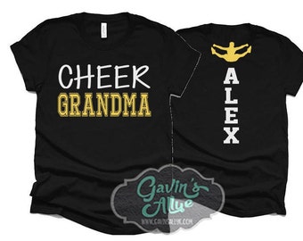 May include: Two black t-shirts. One shirt says "Cheer Grandma" in white and gold lettering. The other shirt has a yellow silhouette of a cheerleader with the name "Alex" spelled vertically in white letters.