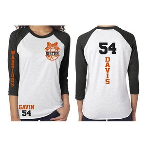 Pu&ograve; includere: Una maglietta da baseball bianca e nera con testo arancione. La parte anteriore della maglietta ha una palla da basket con un fiocco e il testo "Sorella". La parte posteriore della maglietta ha il numero "54" e il testo "Davis". La manica sinistra ha il testo "Warriors" e la parte inferiore della maglietta ha il testo "Gavin 54".