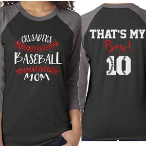 May include: A black and gray baseball mom shirt with red glitter baseball stitching. The shirt says "Crusaders Baseball Mom" with the name "Gavin" on the sleeve. The back of the shirt says "That's My Boy! 10" with a baseball stitched number.