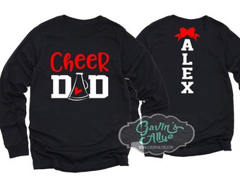 Puede incluir: Dos camisas negras de manga larga con texto blanco. Una camisa dice "Cheer Dad" con un megáfono rojo y un corazón. La otra camisa dice "Alex" con un lazo rojo.