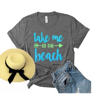 May include: A gray t-shirt with the text "take me to the beach" in blue and green glitter lettering. The shirt is paired with a straw hat and denim shorts.