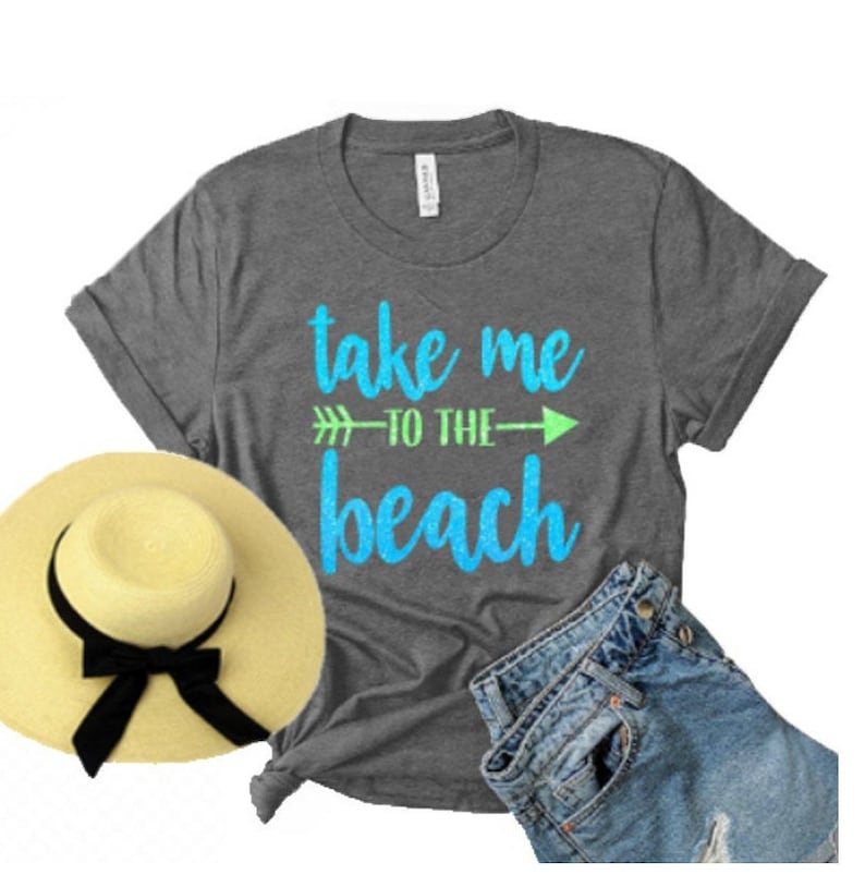 May include: A gray t-shirt with the text "take me to the beach" in blue and green glitter lettering. The shirt is paired with a straw hat and denim shorts.
