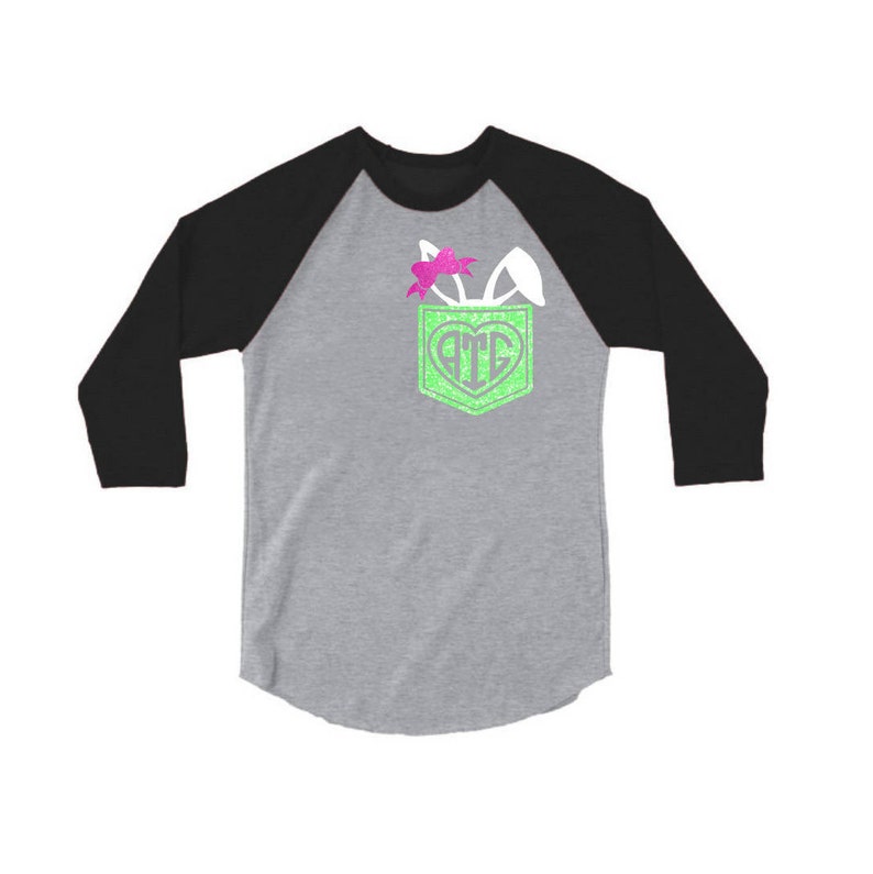 May include: A gray and black baseball tee shirt with a green pocket. The pocket has a white bunny ear sticking out of it with a pink bow. The pocket also has a white heart with the letters "ATE" inside.