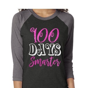 May include: A black and grey baseball tee shirt with pink glitter lettering that reads "100 Days Smarter".