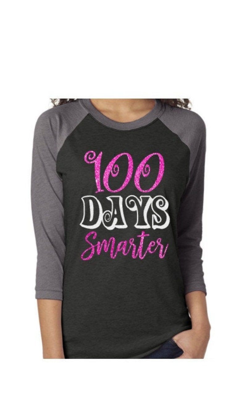 May include: A black and grey baseball tee shirt with pink glitter lettering that reads "100 Days Smarter".