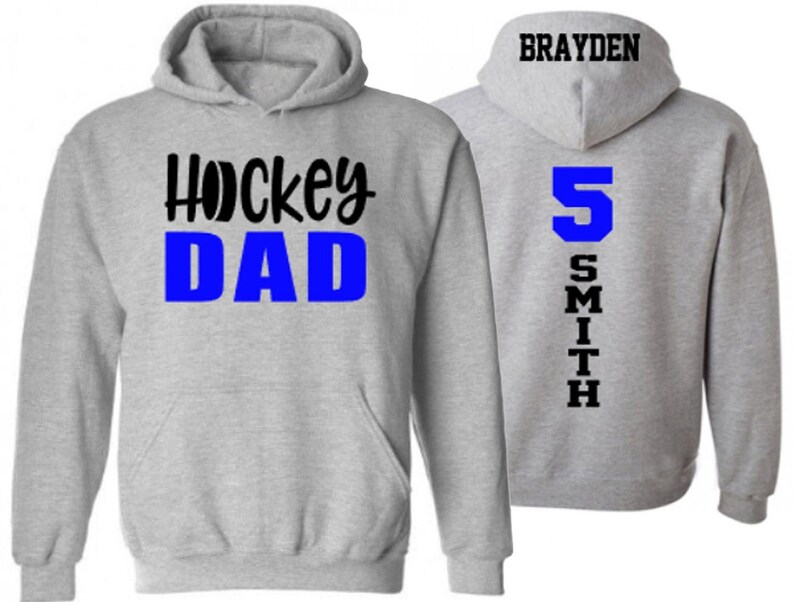 Op de afbeelding: Grijs hoodie met de tekst "Hockey Dad" in zwart en blauw op de voorkant en de tekst "Brayden Smith 5" in zwart en blauw op de achterkant.