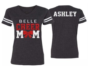 May include: A dark gray t-shirt with white and red glitter lettering that says "Belle Cheer Mom" with a red glitter bow. The back of the shirt has white lettering that says "Ashley".