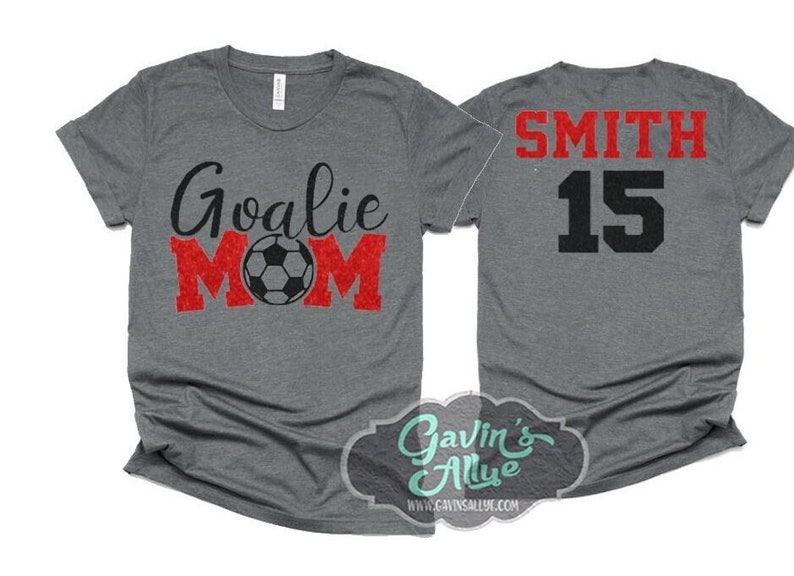 K&ouml;nnte beinhalten: Graues T-Shirt mit dem Text "Goalie Mom" in Schwarz und Rot mit einer Fu&szlig;ballgrafik. Die R&uuml;ckseite des Shirts hat den Text "SMITH 15" in Schwarz und Rot.