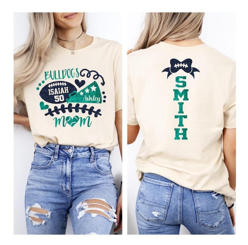 Op de afbeelding: Een cr&egrave;mekleurig t-shirt met een afbeelding op de voorkant van een football, megafoon en de tekst "BULLDOGS", "ISAIAH 50", "ASHLEY" en "MOM" in blauwgroene en marineblauwe glitter. De achterkant toont een football-icoon boven de tekst "SMITH".