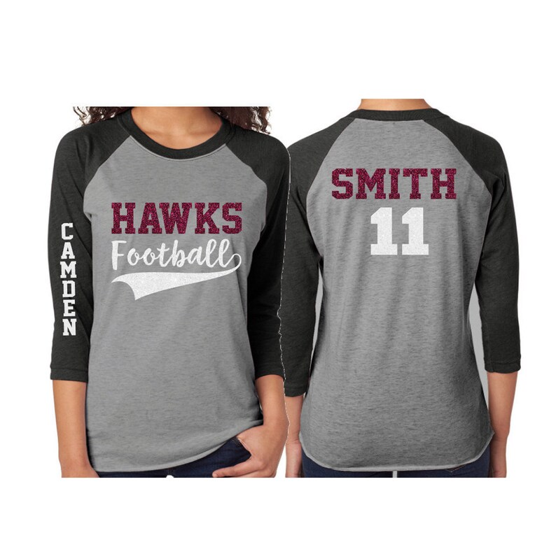 May include: A gray and black baseball jersey with the words "HAWKS Football" in maroon glitter on the front and the name "SMITH" and number "11" in white glitter on the back. The left sleeve has the word "CAMDEN" in white glitter.