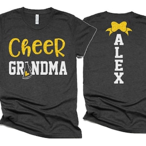 K&ouml;nnte beinhalten: Zwei graue T-Shirts mit goldfarbenem Glitzertext. Das vordere Shirt tr&auml;gt den Schriftzug "Cheer Grandma" mit einem goldfarbenen Glitzermegaphon. Das hintere Shirt hat eine goldfarbene Glitzer Schleife und den Namen "ALEX" in wei&szlig;en Buchstaben.