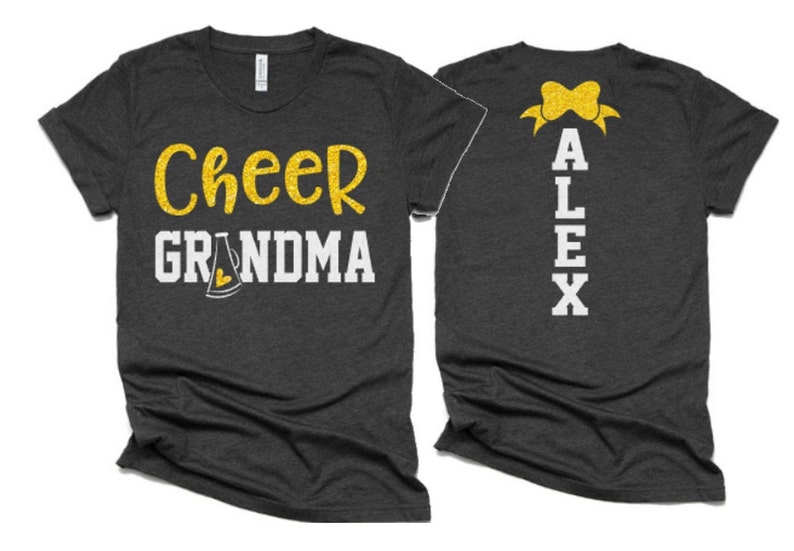 K&ouml;nnte beinhalten: Zwei graue T-Shirts mit goldfarbenem Glitzertext. Das vordere Shirt tr&auml;gt den Schriftzug "Cheer Grandma" mit einem goldfarbenen Glitzermegaphon. Das hintere Shirt hat eine goldfarbene Glitzer Schleife und den Namen "ALEX" in wei&szlig;en Buchstaben.