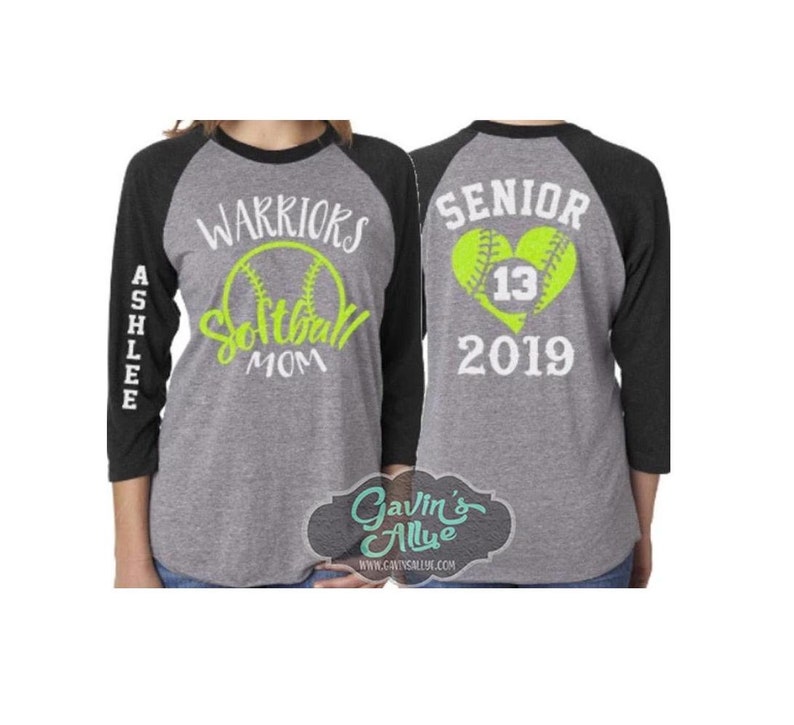 May include: A grey and black baseball t-shirt with the text "WARRIORS Softball MOM" on the front and "ASHLEE" on the left sleeve. The back of the shirt has the text "SENIOR 2019" and a heart with a softball inside. The heart has the number "13" inside.