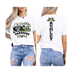 Op de afbeelding: Een wit t-shirt met korte mouwen en een groen-zwarte afbeelding op de voorkant met een football, megafoon en de tekst "FALCONS SYLUS 55 lizzie mom". De achterkant toont de naam "ASHLEY" verticaal met een afbeelding van een groene strik.