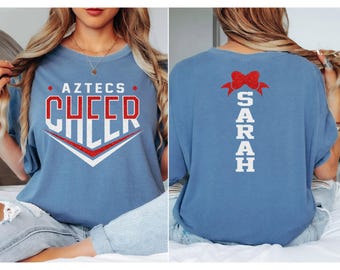 May include: A light blue t-shirt with the words "AZTECS CHEER" in red and white block letters. The back of the shirt has the name "SARAH" in white block letters under a red bow.