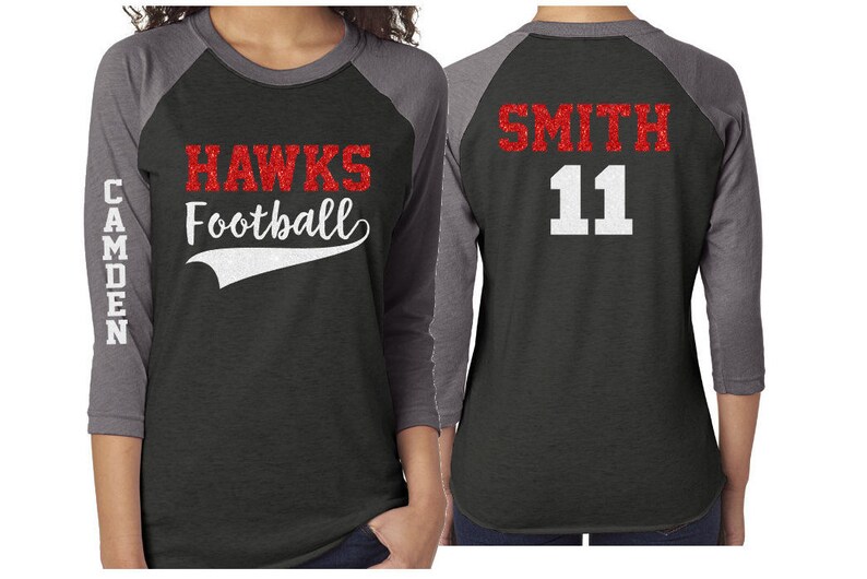 May include: A black and grey baseball jersey with red glitter lettering. The front of the jersey says "HAWKS Football" with a white curved line underneath. The left sleeve says "CAMDEN". The back of the jersey says "SMITH 11" in red glitter lettering.