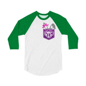 May include: A white and green baseball tee shirt with a pocket on the front. The pocket has a purple glitter heart with the letters "ATG" inside. There are bunny ears and a pink bow coming out of the pocket.