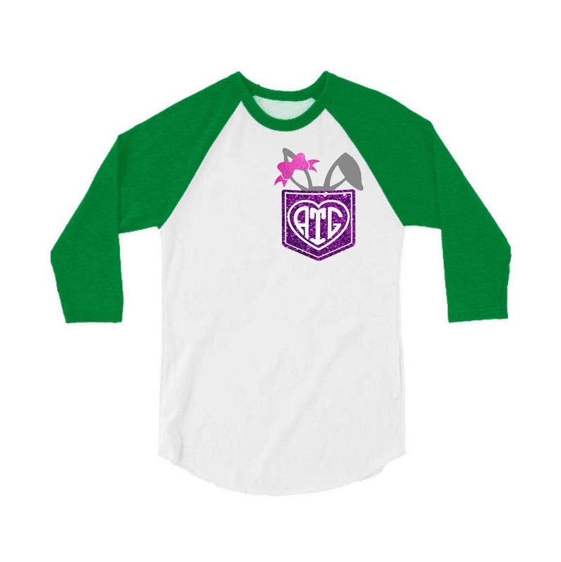 May include: A white and green baseball tee shirt with a pocket on the front. The pocket has a purple glitter heart with the letters "ATG" inside. There are bunny ears and a pink bow coming out of the pocket.