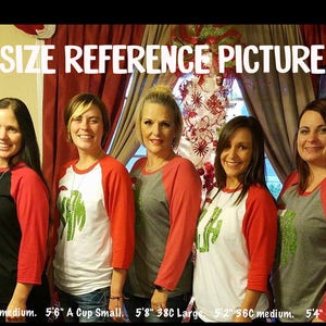 May include: Four women wearing red and grey baseball shirts with green glitter designs. The shirts have the text "SIZE REFERENCE PICTURE" at the top. The women are standing in front of a Christmas tree and a wall with a red curtain. The text at the bottom of the image reads "5'4" 3600 medium. 5'6" A Cup Small, 5'8" 38C Large, 5'2" 36C medium. 5'4" 40C Xlarge."