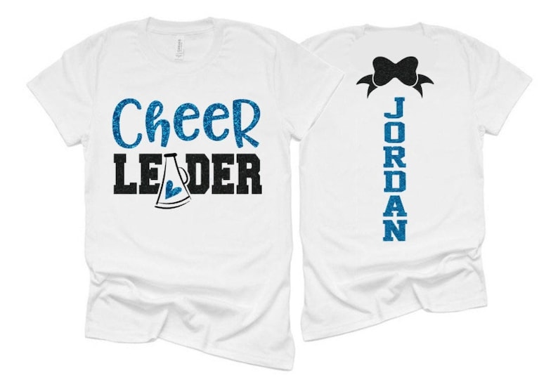 K&ouml;nnte beinhalten: Zwei wei&szlig;e T-Shirts, eines mit dem Text "Cheer Leader" in blauer Glitzer mit einem blauen Glitzer-Megaphon und einem Herzen, und das andere mit dem Text "JORDAN" in blauer Glitzer.