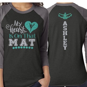 Puede incluir: Una camiseta de b&eacute;isbol negra y gris con un coraz&oacute;n turquesa y una silueta de una bailarina en la parte delantera. El texto "My heart is on that MAT" est&aacute; impreso en la parte delantera de la camiseta. La parte posterior de la camiseta tiene el nombre "ASHLEY" impreso verticalmente en purpurina plateada.