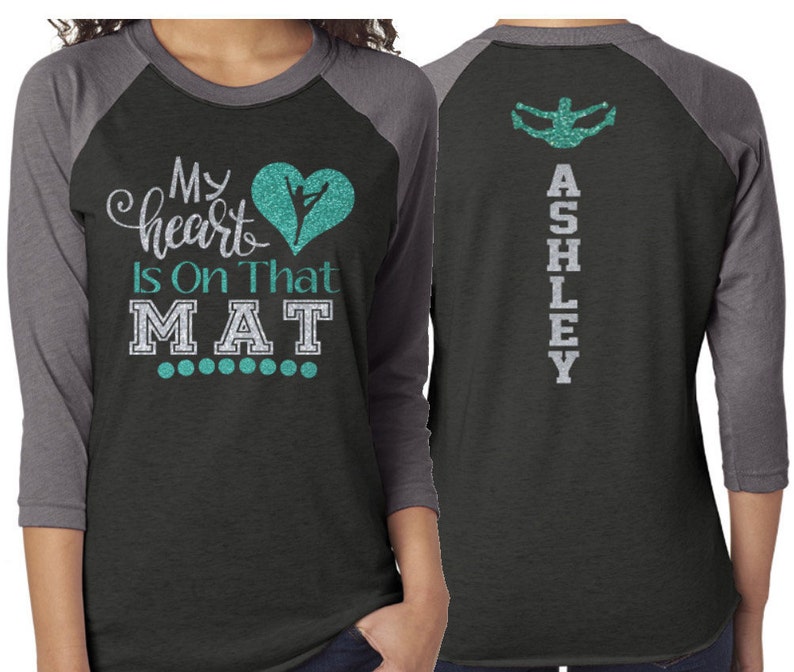 Puede incluir: Una camiseta de b&eacute;isbol negra y gris con un coraz&oacute;n turquesa y una silueta de una bailarina en la parte delantera. El texto "My heart is on that MAT" est&aacute; impreso en la parte delantera de la camiseta. La parte posterior de la camiseta tiene el nombre "ASHLEY" impreso verticalmente en purpurina plateada.
