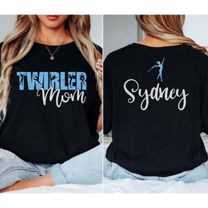May include: Black long-sleeve shirt with the text "TWIRLER Mom" in white script and blue block letters. The back of the shirt has the name "Sydney" in white script and a blue twirler graphic.