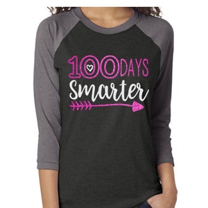 May include: A black and gray raglan t-shirt with pink glitter text that reads "100 Days Smarter" with a small heart and an arrow.