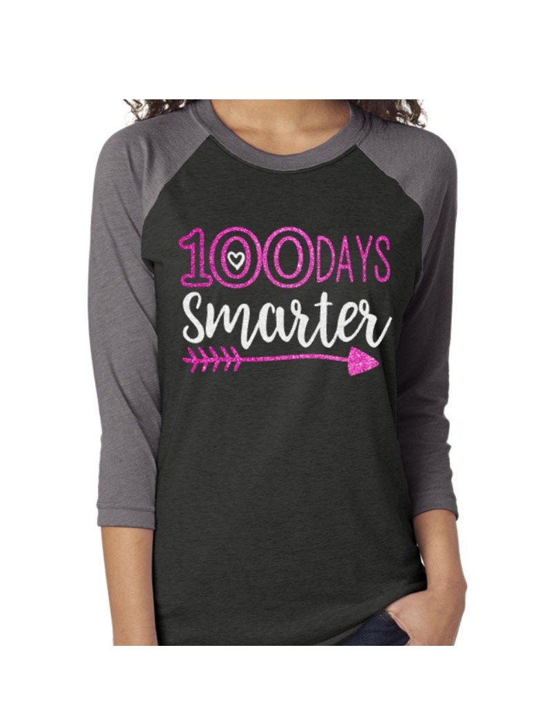 May include: A black and gray raglan t-shirt with pink glitter text that reads "100 Days Smarter" with a small heart and an arrow.