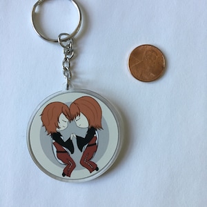 May include: A round keychain with a silver key ring and chain. The keychain features a cartoon illustration of two figures with brown hair, holding hands. They are wearing black jackets and red pants. A penny is next to the keychain.