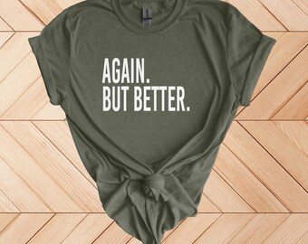 May include: Olive green t-shirt with the text "AGAIN. BUT BETTER." in white block letters. The shirt is knotted at the bottom. The shirt is displayed on a light brown wooden surface.