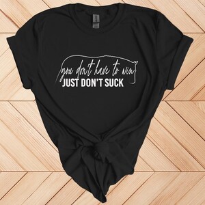 Może przedstawiać: Czarny t-shirt z białym napisem "you don't have to win JUST DON'T SUCK" i konturem konia. Koszulka jest zawiązana na dole. Koszulka jest prezentowana na drewnianej powierzchni.