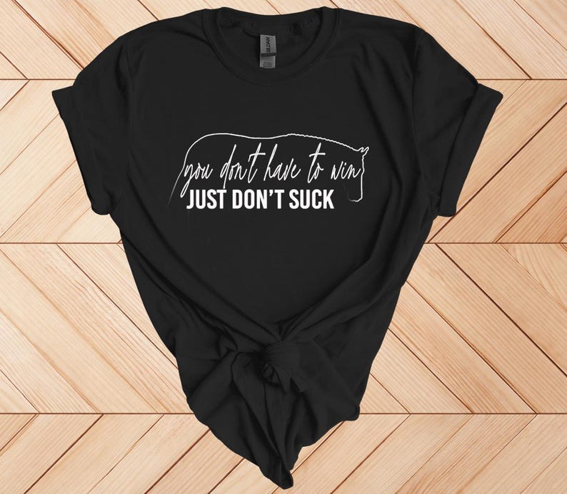 Może przedstawiać: Czarny t-shirt z białym napisem "you don't have to win JUST DON'T SUCK" i konturem konia. Koszulka jest zawiązana na dole. Koszulka jest prezentowana na drewnianej powierzchni.