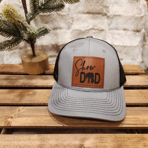 May include: Gray and black trucker hat with a leather patch. The patch features the words "Show Dad" above a silhouette of two elephants. The hat has a curved brim and a mesh back.