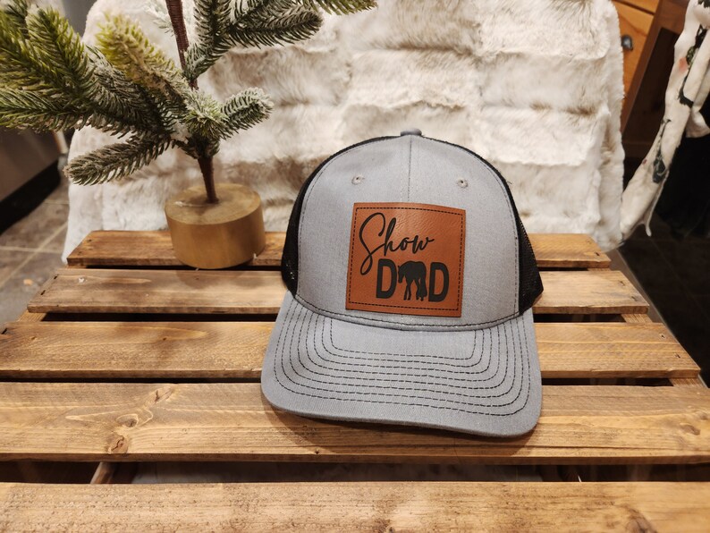 May include: Gray and black trucker hat with a leather patch. The patch features the words "Show Dad" above a silhouette of two elephants. The hat has a curved brim and a mesh back.