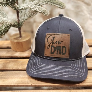 May include: Navy blue trucker hat with a tan mesh back and a brown leather patch. The patch features the words "Show Dad" with a silhouette of a horse. The hat has a curved brim and white stitching.