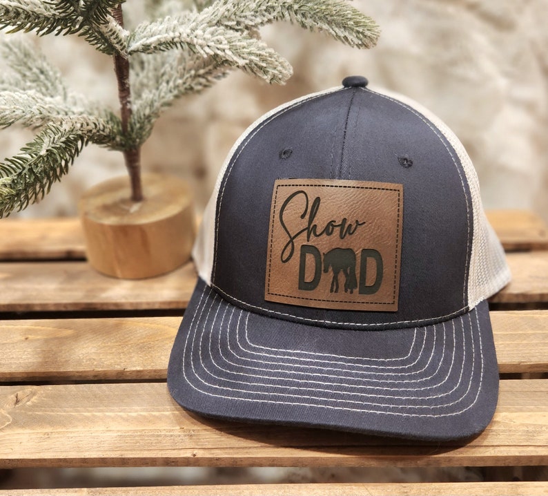 May include: Navy blue trucker hat with a tan mesh back and a brown leather patch. The patch features the words "Show Dad" with a silhouette of a horse. The hat has a curved brim and white stitching.