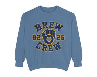 May include: A dusty blue crewneck sweatshirt featuring a graphic print in navy blue and yellow. The design includes the text "BREW CREW" arched around a baseball glove icon with a baseball inside, flanked by the numbers "82" and "26".