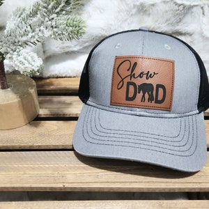 May include: Gray trucker hat with a black mesh back and a brown leather patch. The patch features the words "Show Dad" with a horse silhouette. The hat has a curved brim and a snapback closure.