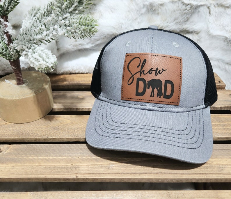 May include: Gray trucker hat with a black mesh back and a brown leather patch. The patch features the words "Show Dad" with a horse silhouette. The hat has a curved brim and a snapback closure.