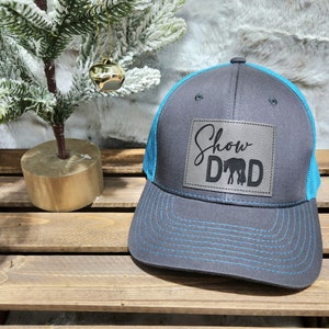 May include: A gray trucker hat with a blue mesh back and brim. The hat features a leather patch with the words "Show Dad" and an image of a horse. The stitching is blue.