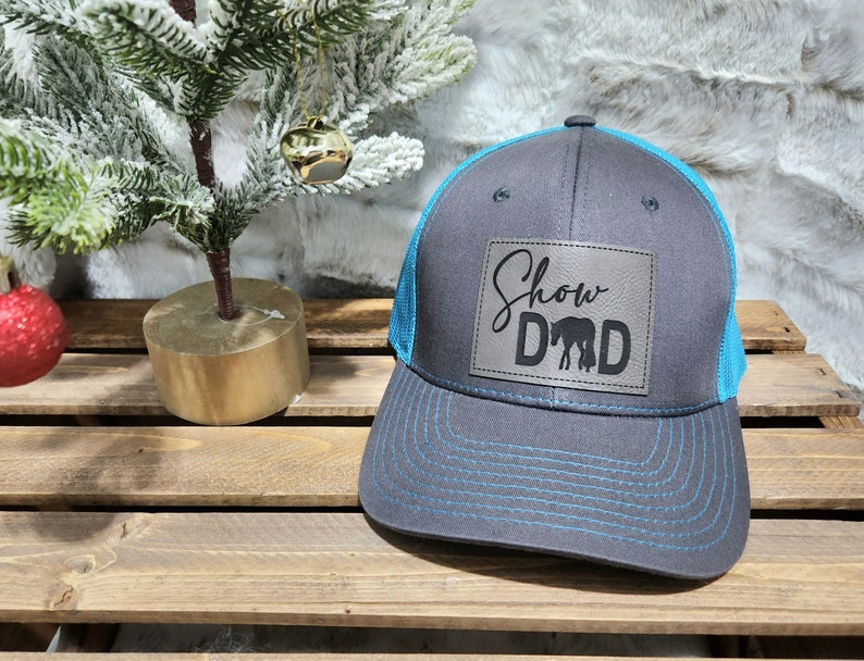 May include: A gray trucker hat with a blue mesh back and brim. The hat features a leather patch with the words "Show Dad" and an image of a horse. The stitching is blue.